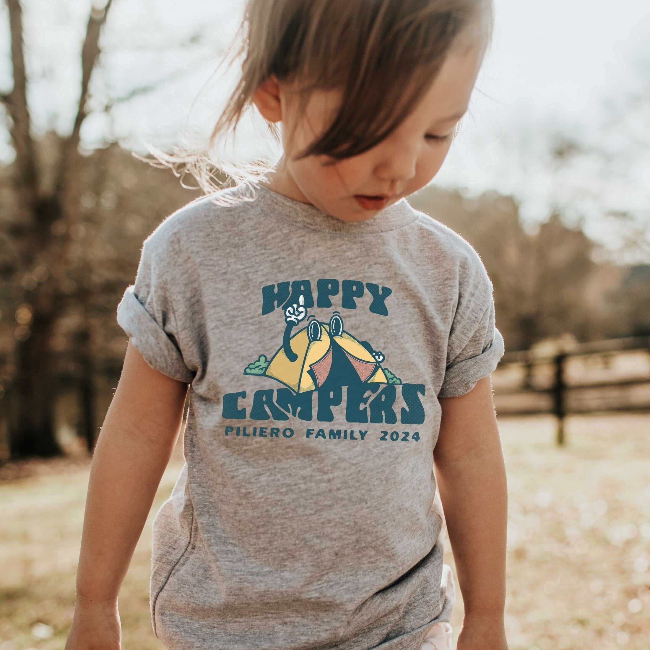 toddler girl wearing custom matching family adventure vacation shirt with retro mascot style Happy Camper cartoon design. Perfect for group camping trips. Shown on gray tshirt