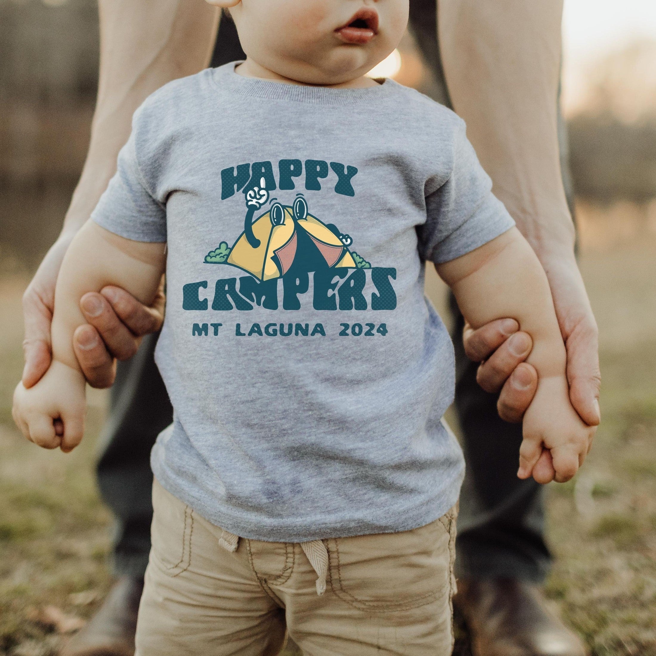 infant wearing custom matching family adventure vacation shirt with retro mascot style Happy Camper cartoon design. Perfect for group camping trips. Shown on gray baby tshirt